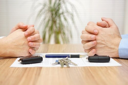 Two pairs of hands folded across a table with an agreement, car key fobs, keys, and pen between them.
