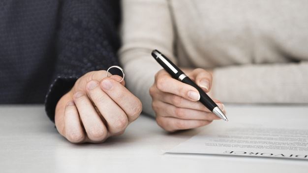 One hand holding a ring and another hand holding a pen signing a divorce agreement.